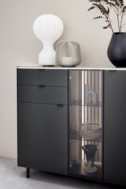 Modernes Highboard in dunklem Design mit Glasvitrine, dekoriert mit einer weißen Lampe, einer grauen Laterne und einer schwarzen Vase. Frontale Perspektive.