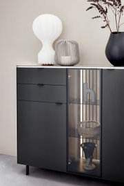 Modernes Highboard in dunklem Design mit Glasvitrine, dekoriert mit einer weißen Lampe, einer grauen Laterne und einer schwarzen Vase. Frontale Perspektive.