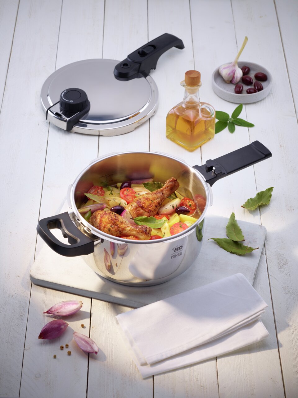 Premium snelkookpan Mistra met geopend deksel, gevuld met verse ingrediënten zoals kippenbouten, tomaten en aardappelen, van bovenaf gefotografeerd op een lichte houten tafel.