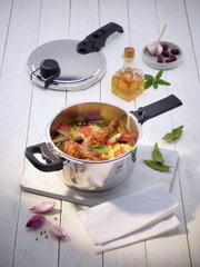 Premium snelkookpan Mistra met geopend deksel, gevuld met verse ingrediënten zoals kippenbouten, tomaten en aardappelen, van bovenaf gefotografeerd op een lichte houten tafel.