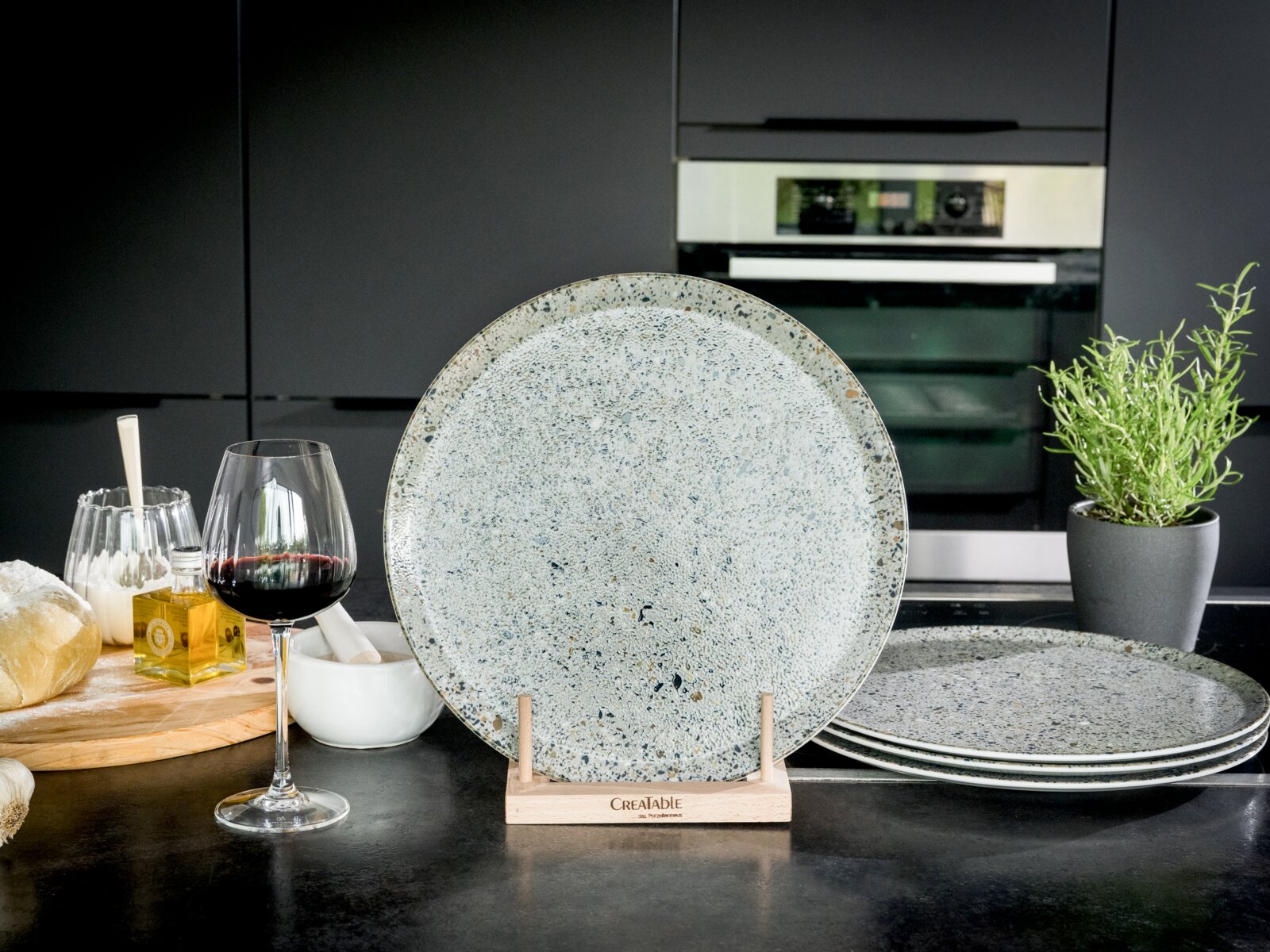 Creatable Pizzateller 4-tlg STONE COLLECTIONF Großer, gesprenkelter Keramik-Speiseteller in Grau/Blau auf Holzständer (Frontalansicht), daneben gestapelte Teller in moderner Küche