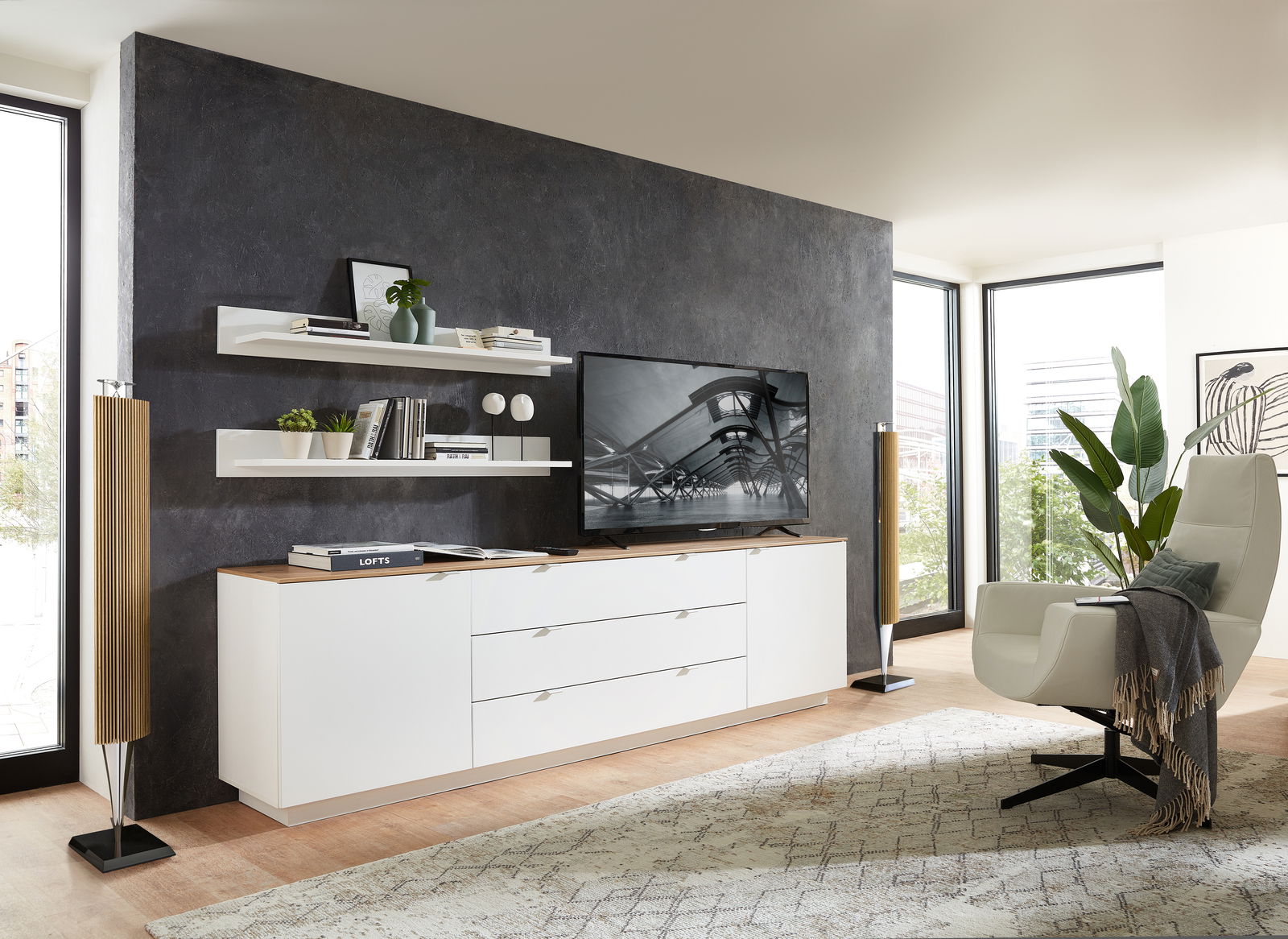 Modernes TV-Sideboard in einem stilvollen Wohnzimmer, seitliche Perspektive. Das Sideboard ist weiß mit einer Holzoberfläche und bietet Platz für einen großen Fernseher. Dekorative Regale und Pflanzen ergänzen das elegante Design.