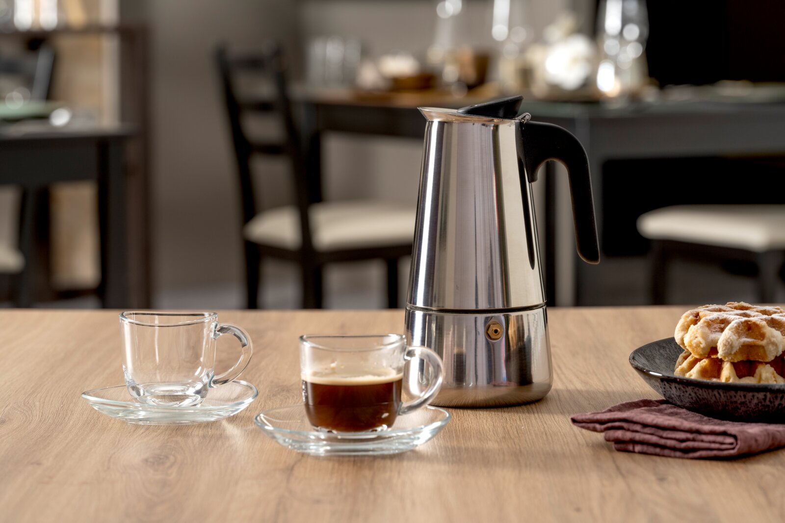 Edelstahl-Espressokocher mit schwarzem Griff auf Holztisch neben Glas-Espressotassen und Gebäck, fotografiert in frontaler Tischperspektive