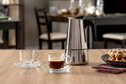 Edelstahl-Espressokocher mit schwarzem Griff auf Holztisch neben Glas-Espressotassen und Gebäck, fotografiert in frontaler Tischperspektive