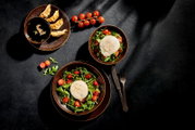 Draufsicht (Flatlay) auf ein stilvolles Food-Arrangement: zwei Schalen Rucola-Tomatensalat mit Burrata, daneben Gyoza mit dunkler Sojasauce, Cherrytomaten am Zweig sowie Messer und Gabel auf dunklem Hintergrund.