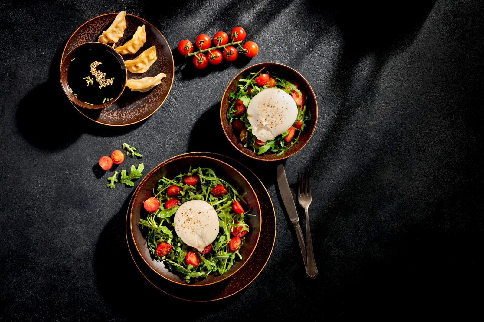 Draufsicht (Flatlay) auf ein stilvolles Food-Arrangement: zwei Schalen Rucola-Tomatensalat mit Burrata, daneben Gyoza mit dunkler Sojasauce, Cherrytomaten am Zweig sowie Messer und Gabel auf dunklem Hintergrund.
