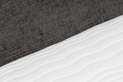 Detailaanzicht van een witte, golvend doorgestikte matras tegen een donkergrijze, geweven bedachterwand, schuin van boven gefotografeerd