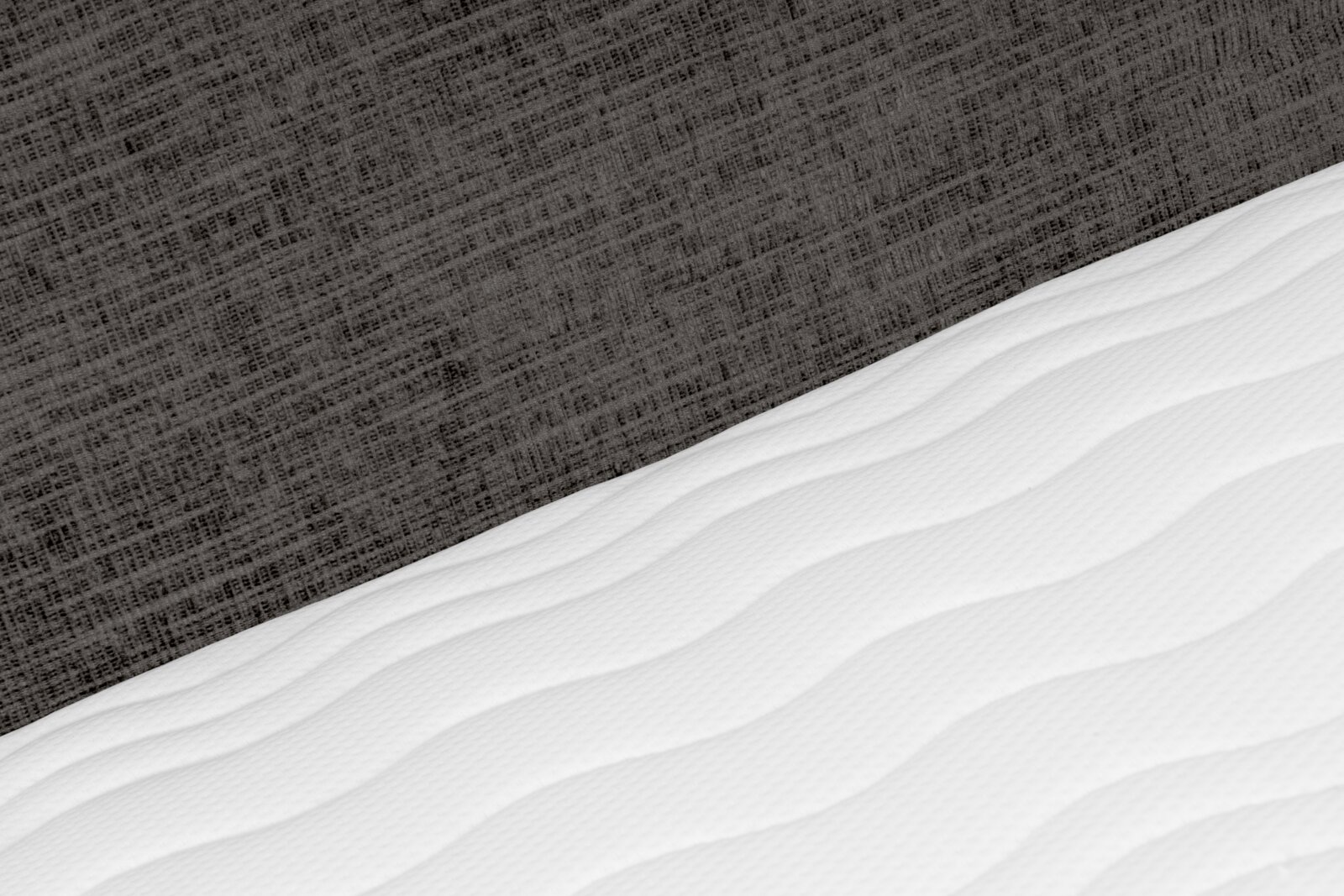 Detailaanzicht van een witte, golvend doorgestikte matras tegen een donkergrijze, geweven bedachterwand, schuin van boven gefotografeerd