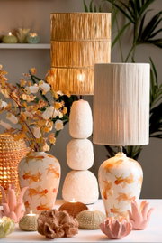 Zwei Tischlampen mit dekorativen Lampenschirmen, eine mit natürlichem Rattan und die andere mit hellem Stoff, auf einem Tisch mit Kerzen und Blumen, seitliche Perspektive.