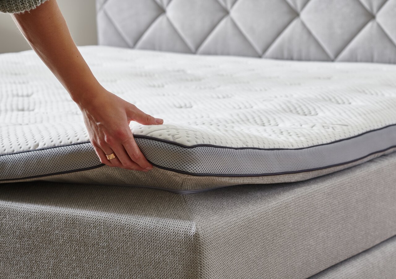 Close-up van een boxspringbed met een hand die het matras optilt om de constructie en het comfort te laten zien.
