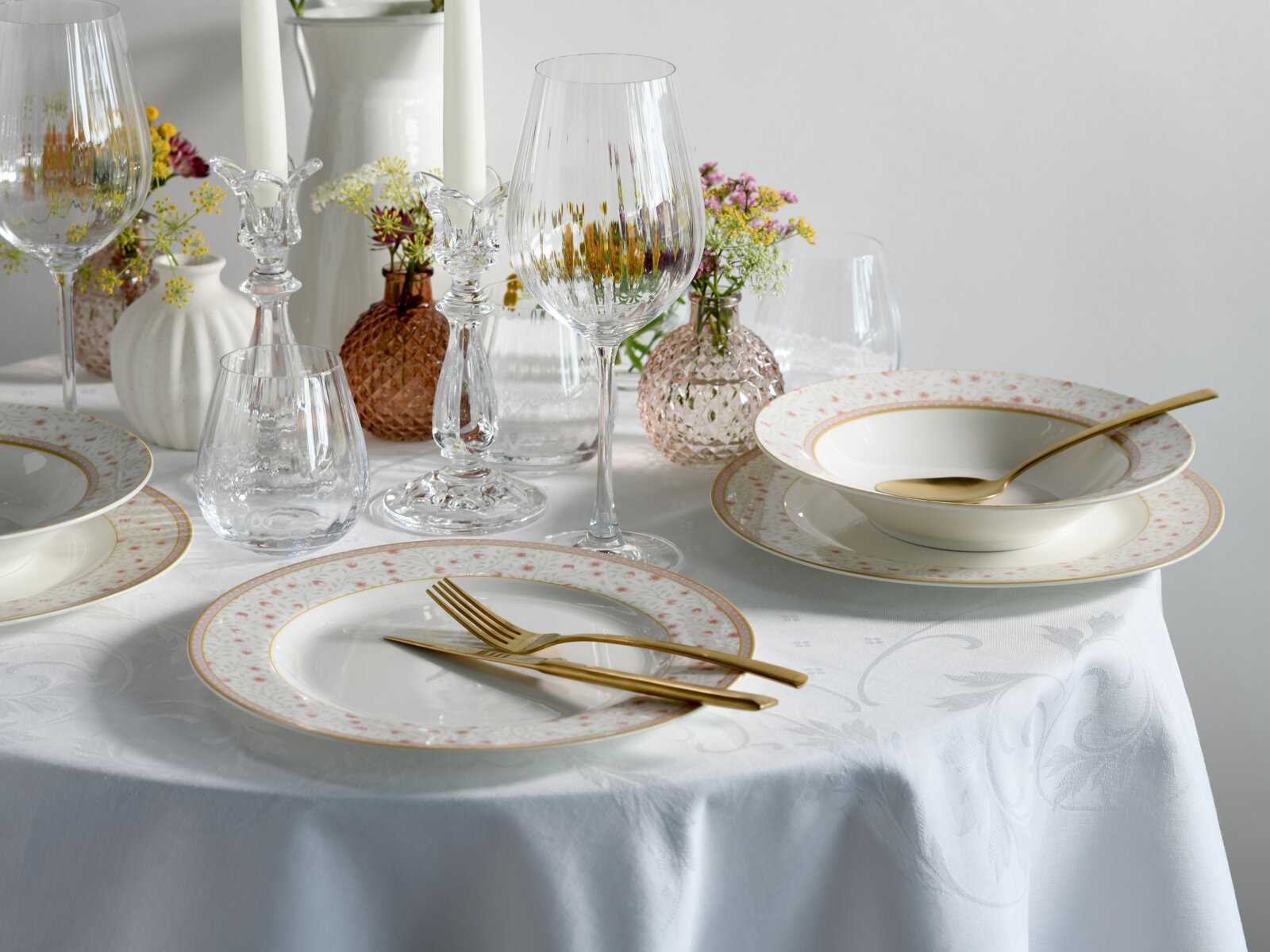 Schick gedeckter Esstisch mit weißem Porzellanservice mit zartem Blumenrand und Goldlinie, goldfarbenem Besteck, Weingläsern und Kerzenleuchtern; Perspektive: leicht erhöhte Frontansicht schräg von vorn.
