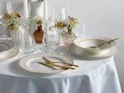 Schick gedeckter Esstisch mit weißem Porzellanservice mit zartem Blumenrand und Goldlinie, goldfarbenem Besteck, Weingläsern und Kerzenleuchtern; Perspektive: leicht erhöhte Frontansicht schräg von vorn.