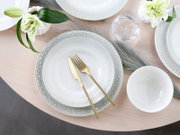 Draufsicht auf eine elegant gedeckte Tafel mit weißen, reliefierten Tellern mit hellgrauem Muster, goldfarbenem Messer und Gabel, Wasserglas, weißer Schale und weißen Blumen auf hellem Holztisch.