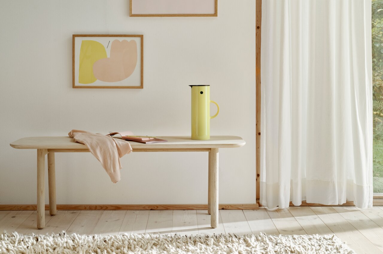 Gelbe EM77 Isolierkanne, 1L, auf einem Holztisch in einem hellen Raum mit moderner Kunst an der Wand, seitliche Perspektive.
