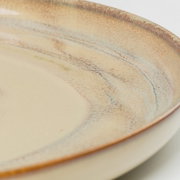 Nahaufnahme eines runden Keramiktellers in Beige mit braun-grauer, gesprenkelter Glasur und feinen Linien, perspektivisch leicht schräg von oben fotografiert.