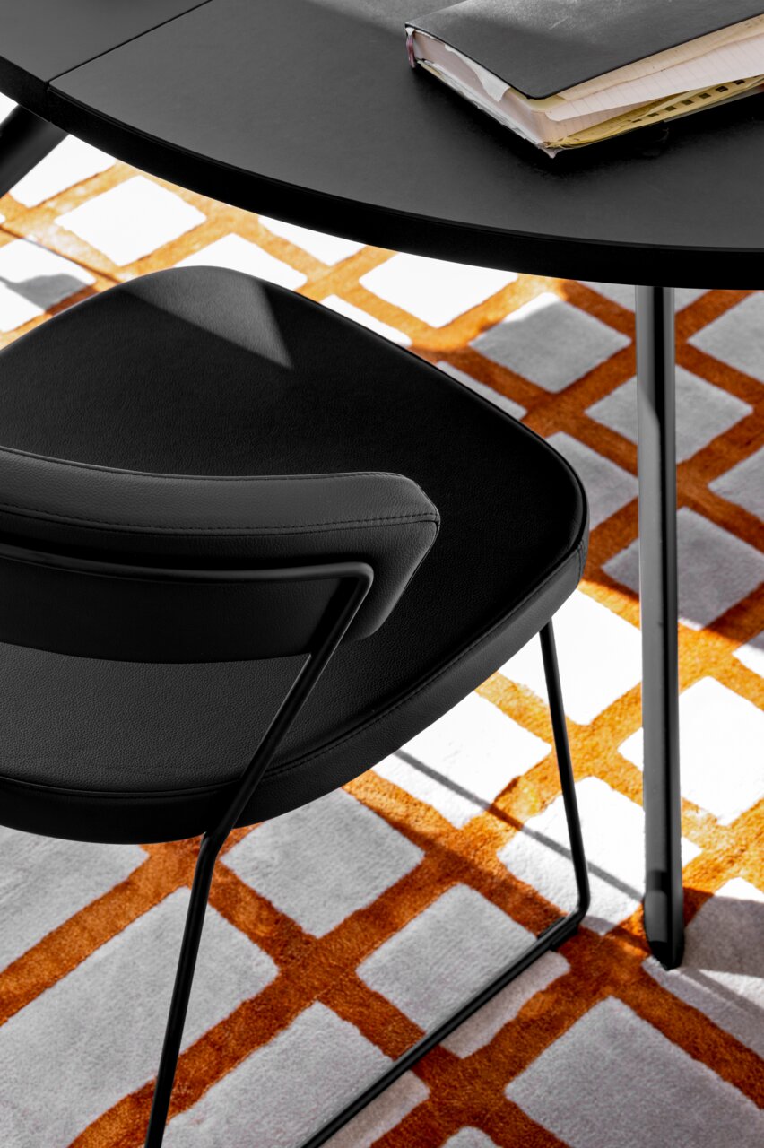 Moderne zwarte stoel met metalen frame, gefotografeerd van bovenaf, op een tapijt met een oranje-wit patroon.