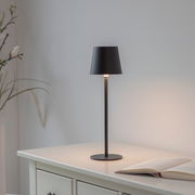 Schwarze Akku-Tischleuchte BOTERIA auf einem weißen Tisch, seitliche Perspektive, mit einem geöffneten Buch daneben.