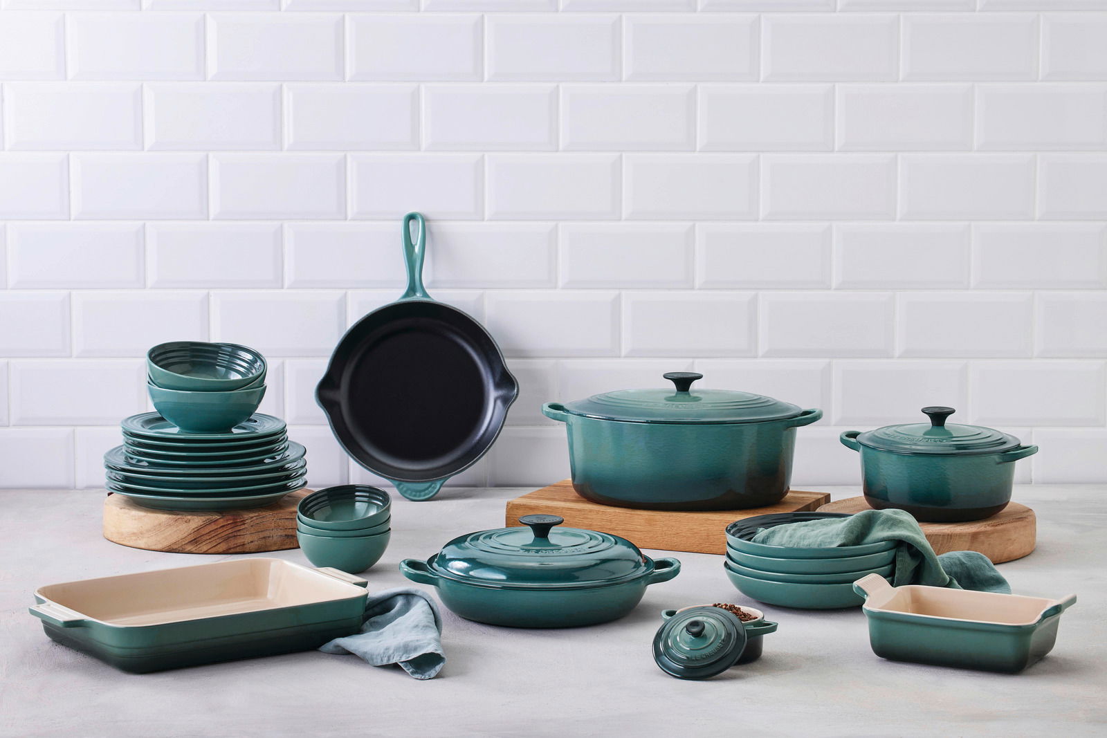 LE CREUSET Gourmet-Profitopf 26cm Tradition OCEAN Verschiedene Küchenutensilien in der Farbe Ocean, darunter Bräter, Pfannen und Auflaufformen, auf einer Arbeitsfläche vor einer weißen Fliesenwand. Perspektive von vorne.