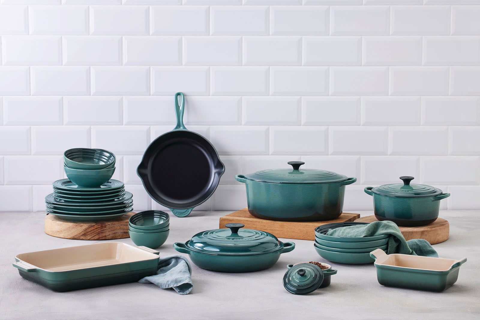 LE CREUSET Gourmet-Profitopf 26cm Tradition OCEAN Verschiedene Küchenutensilien in der Farbe Ocean, darunter Bräter, Pfannen und Auflaufformen, auf einer Arbeitsfläche vor einer weißen Fliesenwand. Perspektive von vorne.