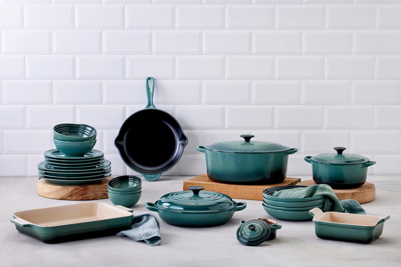 LE CREUSET Gourmet-Profitopf 26cm Tradition OCEAN Verschiedene Küchenutensilien in der Farbe Ocean, darunter Bräter, Pfannen und Auflaufformen, auf einer Arbeitsfläche vor einer weißen Fliesenwand. Perspektive von vorne.