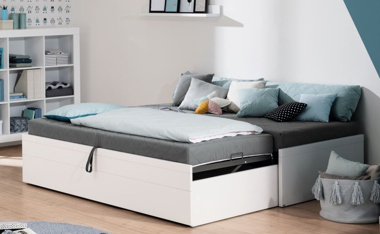 Moderne Flynn-Funktionsliege in einem hellen Raum, seitliche Perspektive, mit grauer Matratze und mehreren Kissen in verschiedenen Farben.