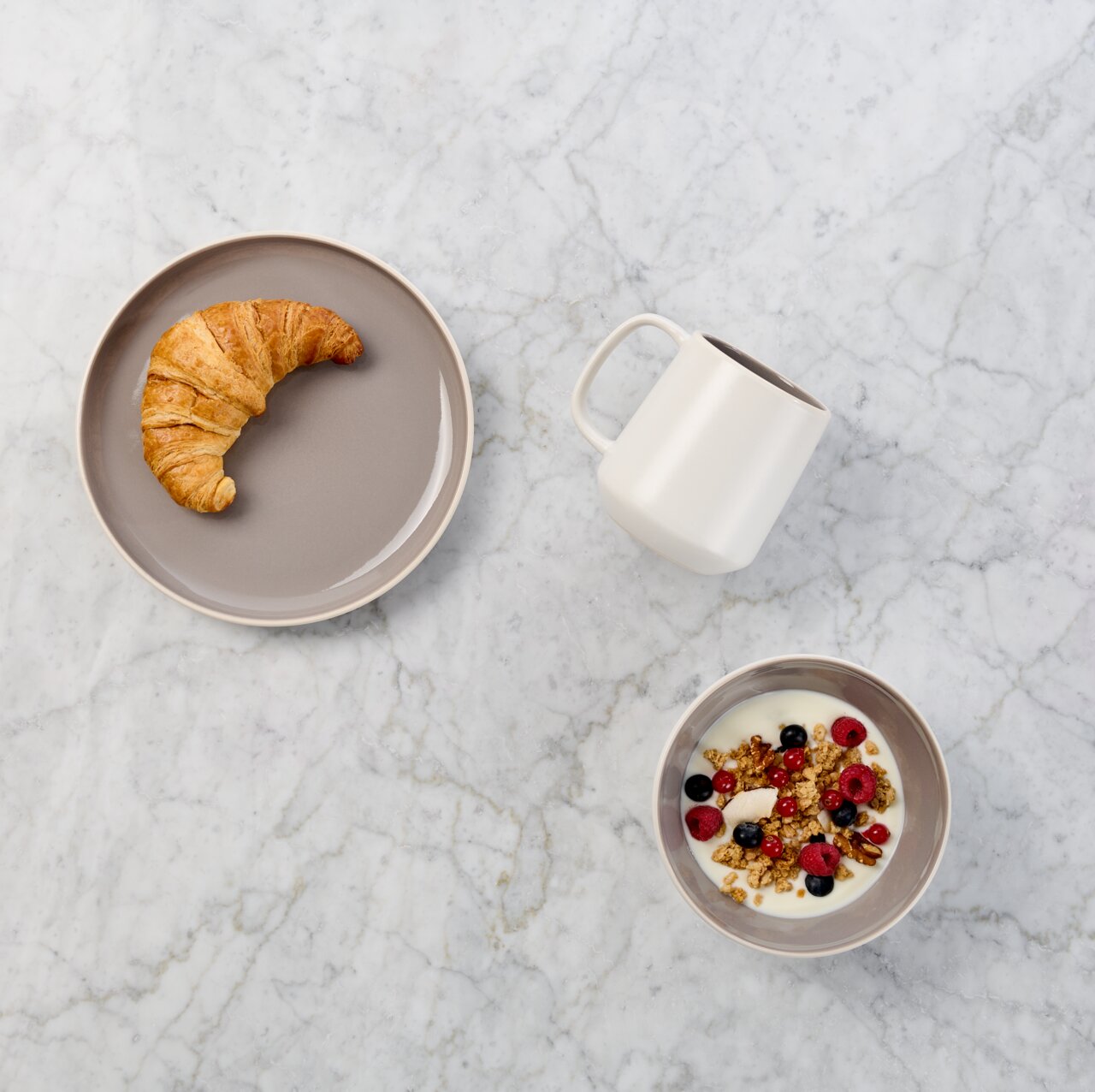 Draufsicht auf ein Frühstücks-Arrangement auf heller Marmorplatte: Croissant auf grauem Teller, weiße Tasse und Schale mit Joghurt, Granola und Beeren.