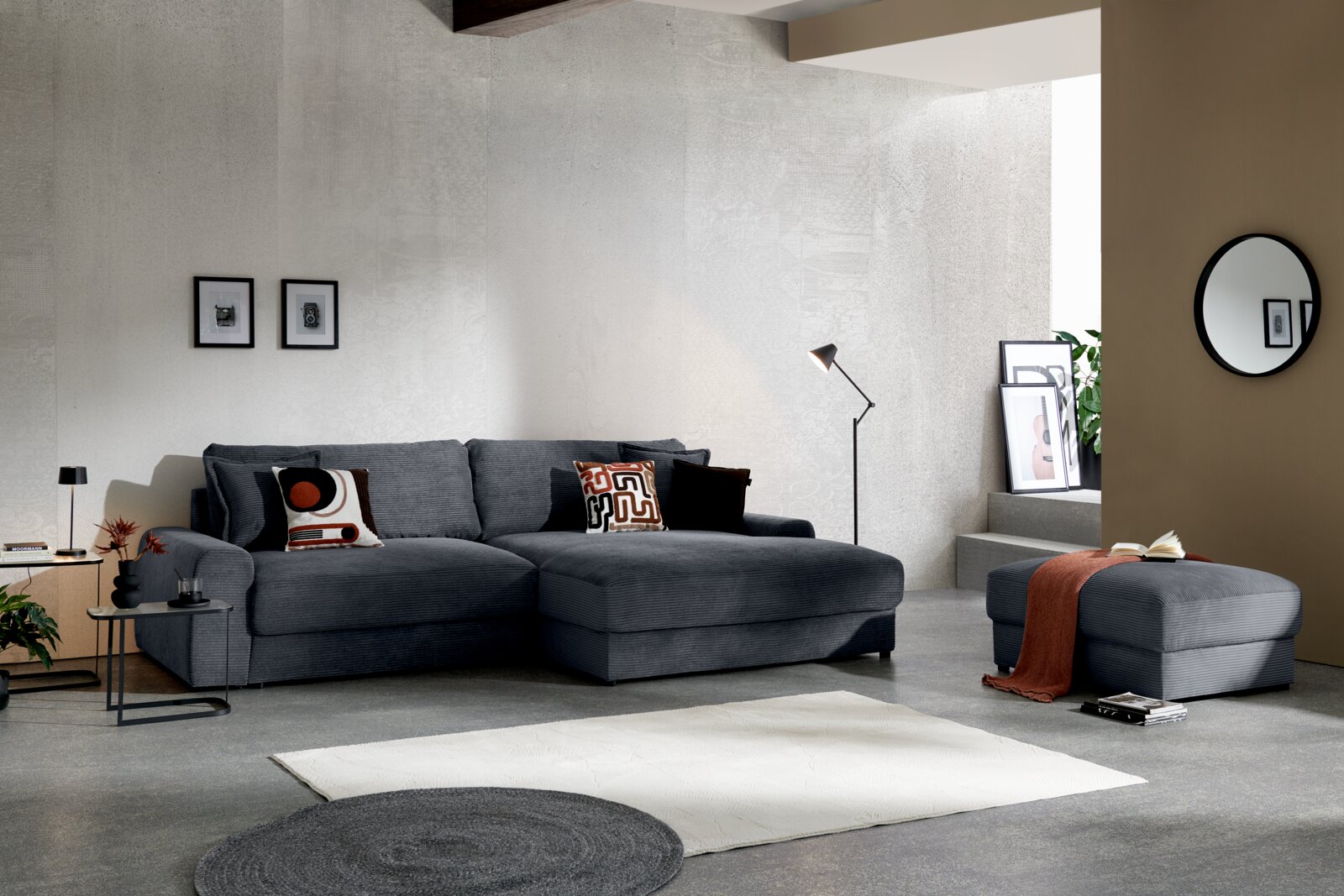Modernes Wohnzimmer mit grauem Stoff-Ecksofa mit rechter Recamiere und passendem Hocker, dekoriert mit Kissen und Decke; Perspektive: frontale, leicht seitliche Ansicht auf Sofa und Hocker.