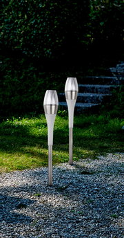 Zwei moderne Solarleuchten aus Edelstahl und Kunststoff, aufgestellt in einem Garten mit Kiesweg und Treppen im Hintergrund, seitliche Perspektive.