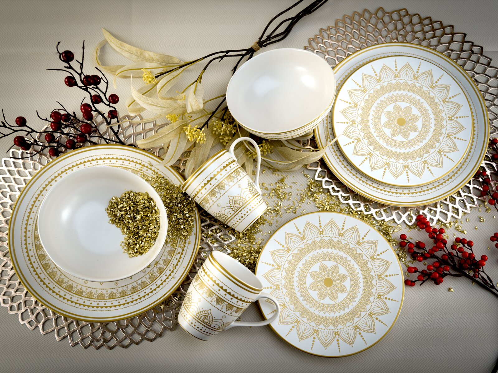 Draufsicht auf elegantes weißes Geschirrset mit goldfarbenem Mandala-Dekor (Teller, Schalen und Tassen) dekorativ arrangiert auf Tischunterlage mit Beerenzweigen.