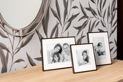 Drei schwarze Aluminium-Bilderrahmen auf einem Holztisch, vor einer Wand mit Blattmuster, aus seitlicher Perspektive fotografiert.