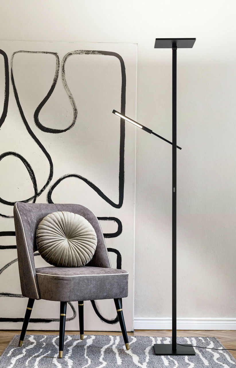 Fabas Luce Stehleuchte IDEAL Schwarze Stehleuchte in modernem Design, neben einem grauen Stuhl mit runder Kissenlehne, vor einer abstrakten Wandkunst. Seitliche Perspektive.