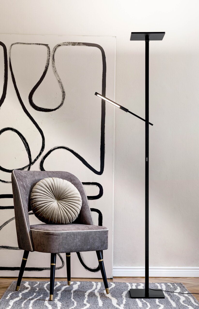 Schwarze Stehleuchte in modernem Design, neben einem grauen Stuhl mit runder Kissenlehne, vor einer abstrakten Wandkunst. Seitliche Perspektive.