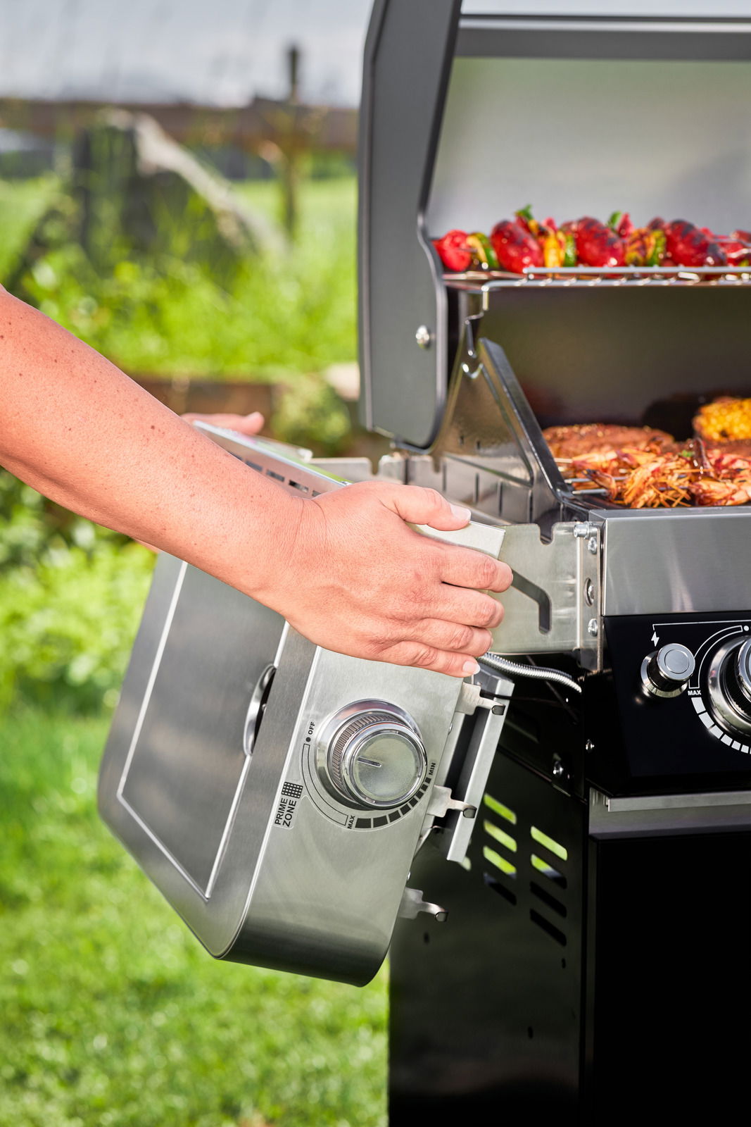 Nahaufnahme einer BBQ-Station Videro G4-S mit geöffneter Haube, auf der Grillgut zu sehen ist. Eine Hand bedient den seitlichen Brenner. Perspektive von der Seite.