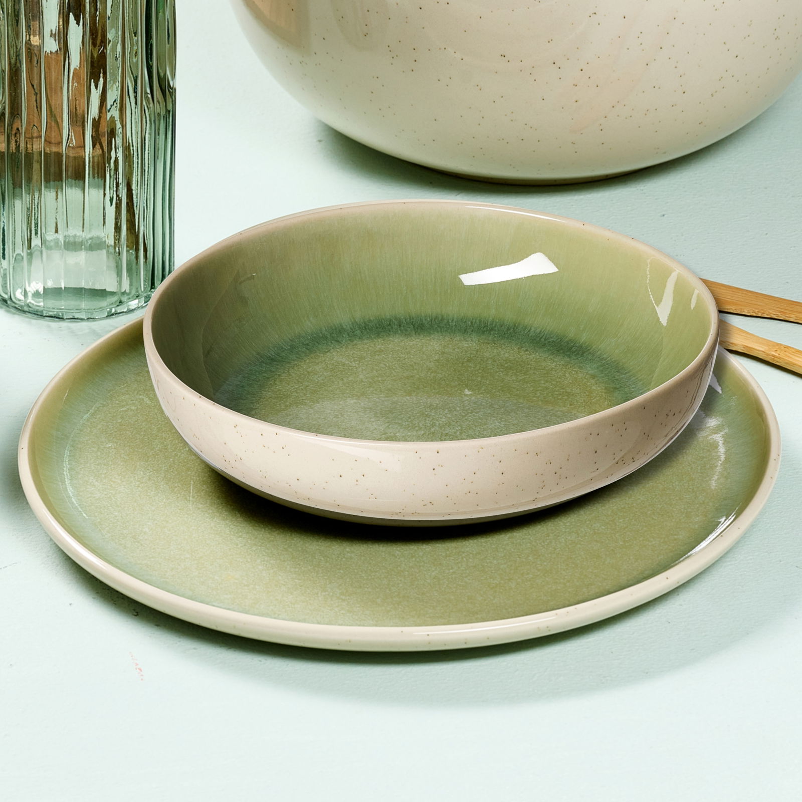 Ritzenhoff & Breker Tafelset 8-tlg. SKOG Nahaufnahme eines hellgrünen Tafelsets, bestehend aus einem flachen Teller und einer Schale, auf einem hellen Untergrund. Im Hintergrund sind ein Glas und ein Teil einer großen Schüssel zu sehen.