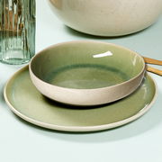 Nahaufnahme eines hellgrünen Tafelsets, bestehend aus einem flachen Teller und einer Schale, auf einem hellen Untergrund. Im Hintergrund sind ein Glas und ein Teil einer großen Schüssel zu sehen.