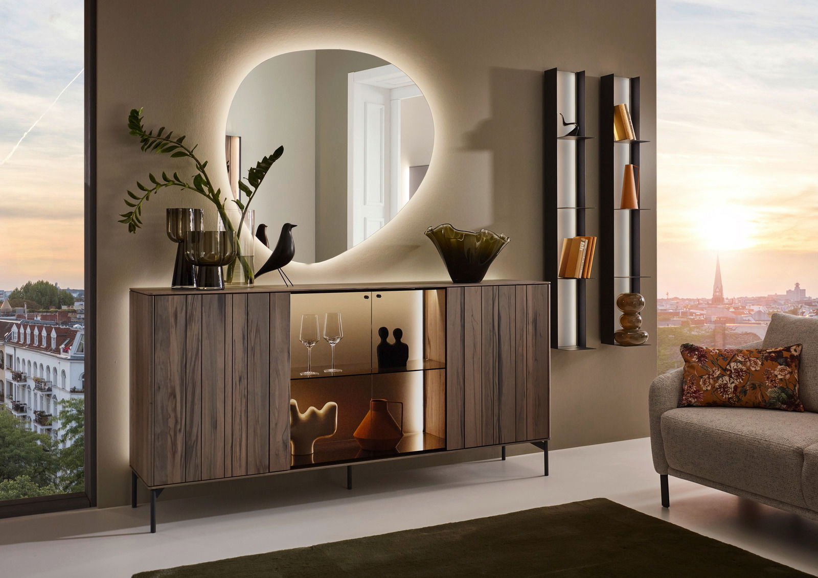 Johann Jakob Wandspiegel LANDO Moderner Wandspiegel mit organischer Form, beleuchtet, über einem stilvollen Sideboard in einem eleganten Wohnzimmer mit Blick auf die Stadt bei Sonnenuntergang.