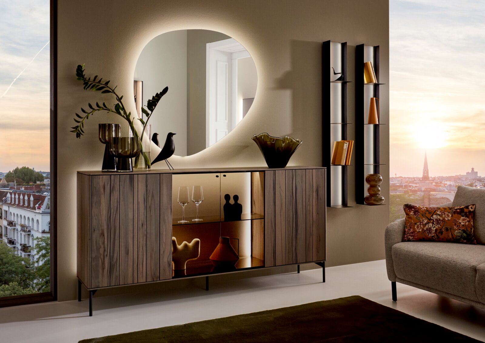 Moderne wandspiegel met organische vorm, verlicht, boven een stijlvol dressoir in een elegante woonkamer met uitzicht op de stad bij zonsondergang.
