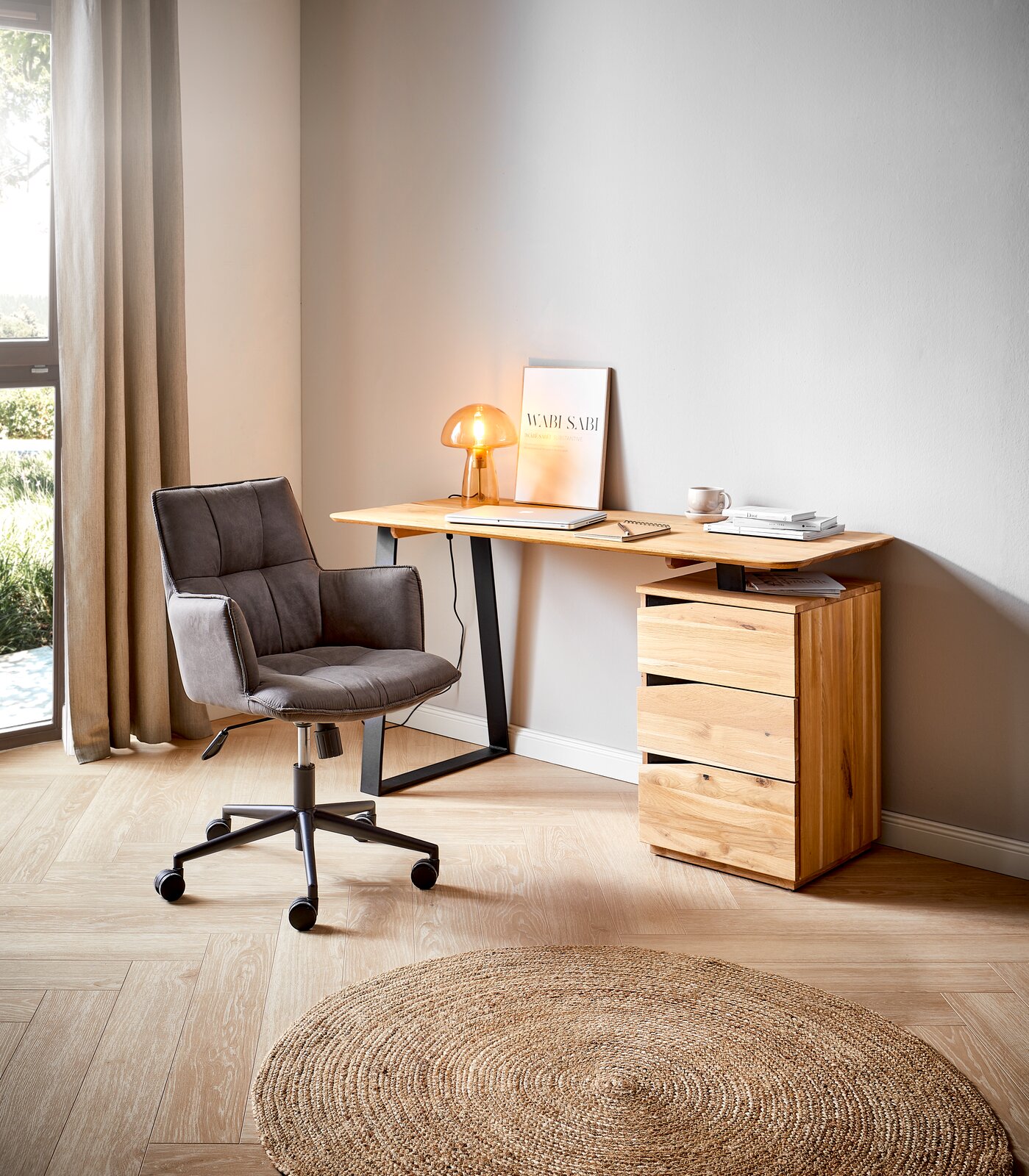Moderner Schreibtisch aus Holz mit schwarzem Metallgestell, seitlich von einem grauen Bürostuhl und einer kleinen Tischlampe begleitet, in einem hellen Raum mit Holzboden und natürlichem Licht.