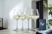 Vier elegante Weißweingläser, gefüllt mit Weißwein, auf einem Tisch arrangiert. Die Perspektive zeigt die Gläser von der Seite, mit einem dekorativen Element im Hintergrund.
