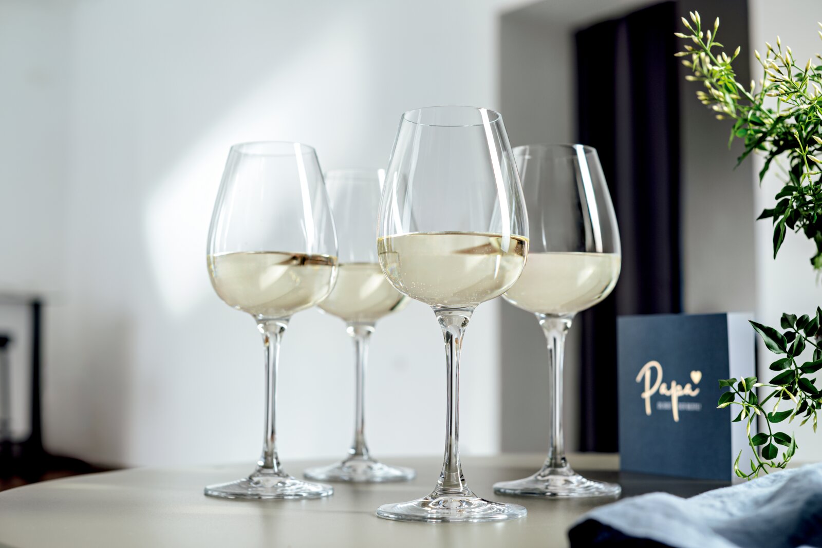 Vier elegante Weißweingläser, gefüllt mit Weißwein, auf einem Tisch arrangiert. Die Perspektive zeigt die Gläser von der Seite, mit einem dekorativen Element im Hintergrund.