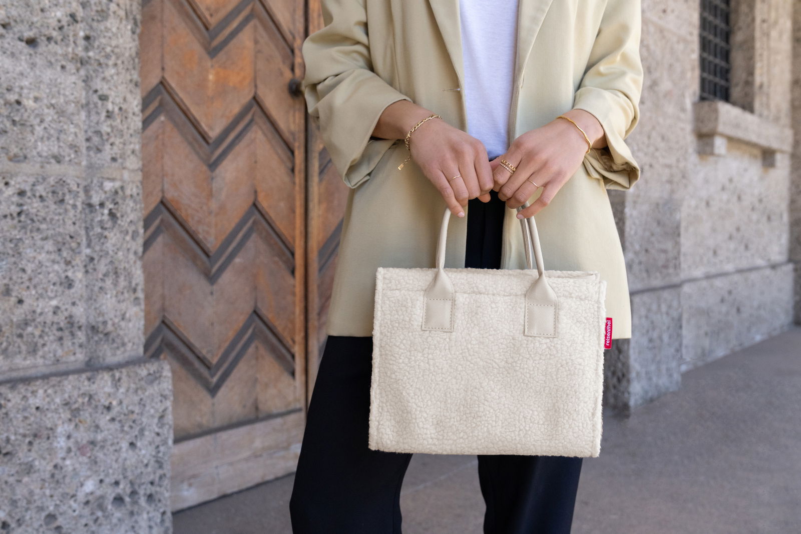 Frau hält eine kleine, cremefarbene Shopper-Tasche aus weichem Material vor einem Holztor. Perspektive: Frontalansicht.