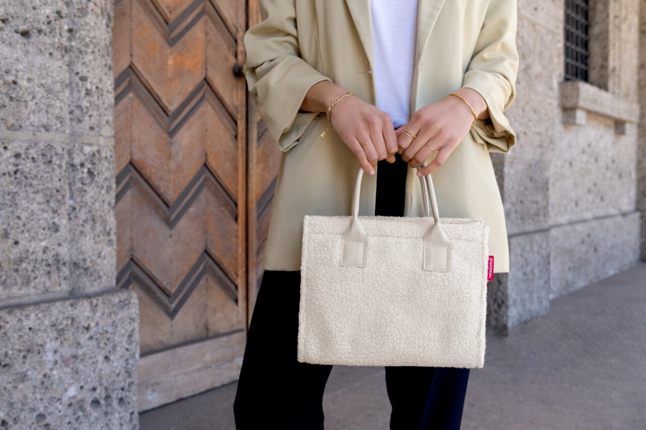 Frau hält eine kleine, cremefarbene Shopper-Tasche aus weichem Material vor einem Holztor. Perspektive: Frontalansicht.