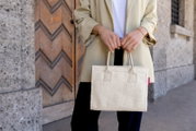 Frau hält eine kleine, cremefarbene Shopper-Tasche aus weichem Material vor einem Holztor. Perspektive: Frontalansicht.