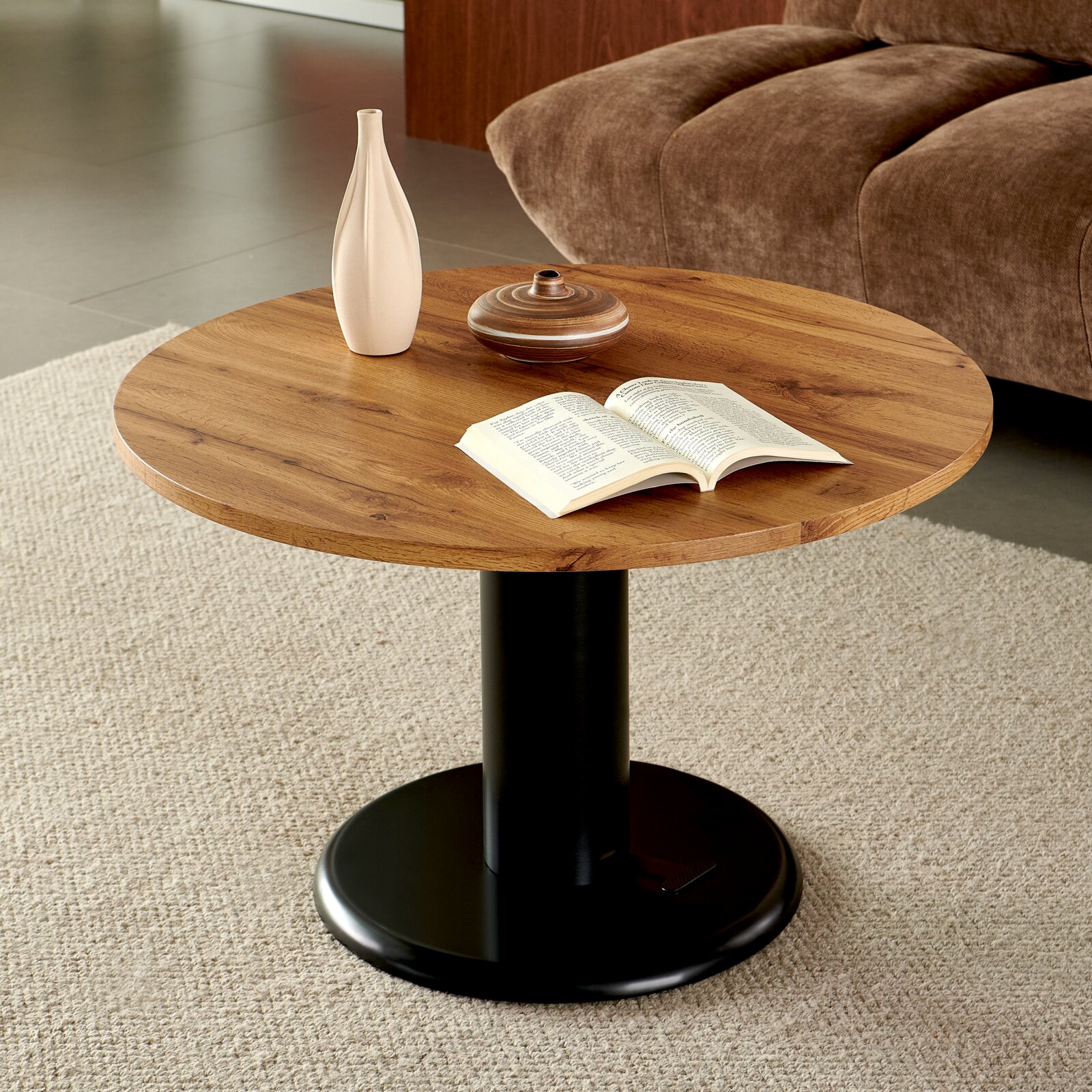 Runder Couchtisch mit Holzplatte und schwarzem Mittelfuß auf Teppich, darauf Vase, kleine Schale und offenes Buch perspektivische Ansicht leicht von oben im Wohnzimmer neben Sofa