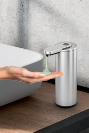 Edelstahl-Seifenspender mit Sensor gibt grünes Flüssigseifen-Gel in eine ausgestreckte Hand neben einem Waschbecken ab, seitliche Nahaufnahme aus leicht erhöhter Perspektive