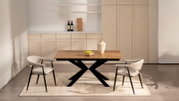 Moderner Esstisch aus Holz mit schwarzem X-förmigem Gestell, dekoriert mit Vase und Schale Trauben, flankiert von zwei gepolsterten Stühlen; frontale Perspektive auf das Set in hellem Wohnraum.