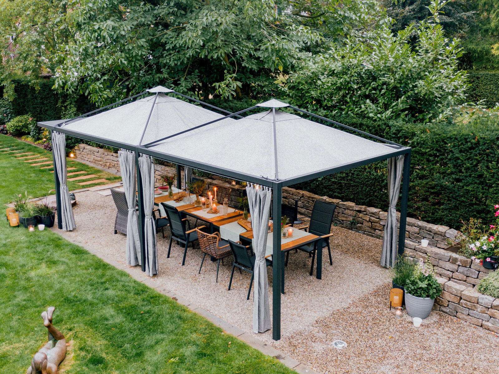Siena Garden Pagode Erweiterung Luftaufnahme einer eleganten Gartenpagode mit Vorhängen, darunter ein Esstisch mit Stühlen, umgeben von grüner Vegetation.