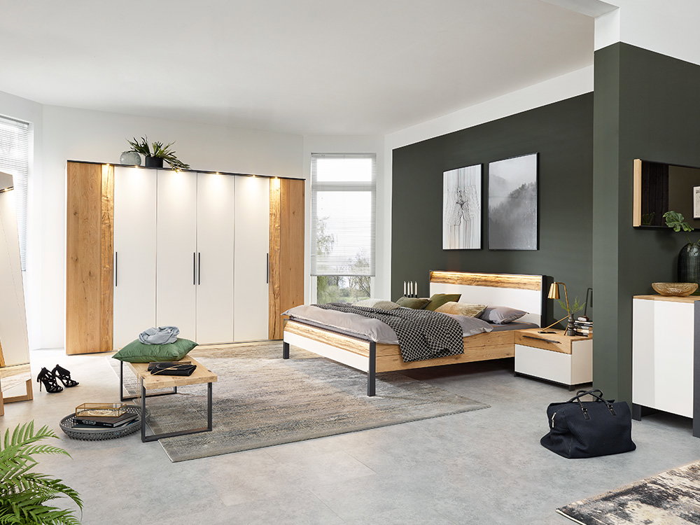 Das Bild zeigt ein modernes Schlafzimmer. Das Bett ist aus Holz mit einem schlichten Design. Daneben stehen Nachttisch mit Lampen. Im Hintergrund sieht man einen großen Kleiderschrank mit Holz- und weißen Elementen. Die Wände sind teilweise dunkelgrün ges