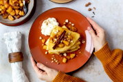 Draufsicht auf einen orangefarbenen Teller mit gefalteten Crêpes, getoppt mit Apfelstückchen, gehackten Nüssen und Zimtstange, daneben ein Klecks Schlagsahne; Hände halten den Teller über hellem Tisch.