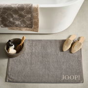 Graue Badematte mit 'JOOP!' Schriftzug, platziert vor einer Badewanne, umgeben von einem Korb mit Pflegeprodukten und beigen Hausschuhen, aus der Vogelperspektive.